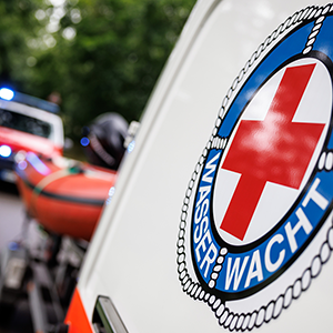 Nahaufnahme des Wasserwacht-Logos auf weißem Fahrzeug, im unscharfen Hintergrund ein Rettungsboot und Blaulichter.